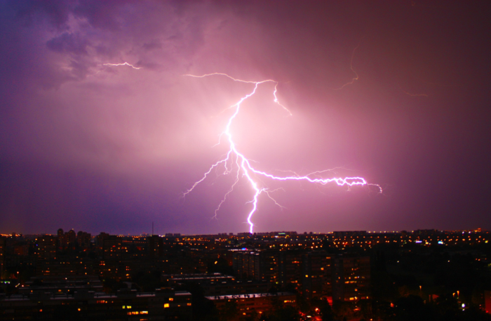 A violet sky is lit by a gigantic thunderbolt striking a city. 