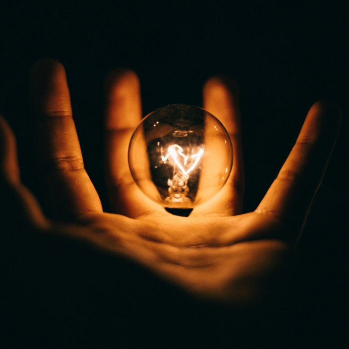 A light bulb glows in the darkness illuminating a hand that almost touches it like the bulb is floating above the hand. 
